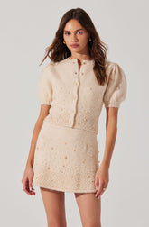 The outfit consists of a cream-colored, short-sleeved, knitted cardigan with puffed sleeves and small decorative pearls. The matching mini skirt is also cream-colored and features a similar pearl embellishment, creating a coordinated, textured look.